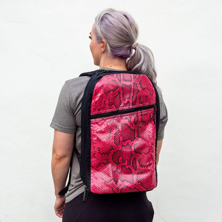 A person with light purple hair tied back is sporting a light gray t-shirt and carrying the GORUCK Bullet Ruck Heritage in Pink Python. The background features a plain, light-colored wall, which accentuates their modern style with vintage inspiration reminiscent of the Martexin Wax finish.