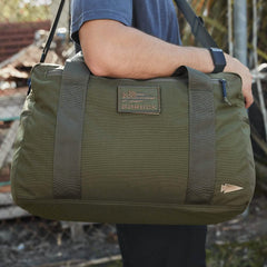 A person in a blue shirt carries an olive green Kit Bag from GORUCK, similar to a Special Forces Flight Bag, featuring sturdy straps. The bag displays the GORUCK patch with an arrow design. Outdoor elements like foliage and fencing provide the backdrop to this rugged scene.
