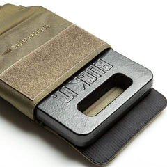 A weighted ruck plate is partially inserted into the tan, robust Ruck Plate Carrier 3.0 from GORUCK, secured with a Velcro flap. Crafted from 1000D CORDURA, this durable carrier is ideal for fitness or military training and offers ergonomic lumbar support. The black plate features an embossed rectangular hole with the text RUCK PL.