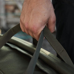 A close-up of a person's hand gripping the handle of a GORUCK Kit Bag, available in green and reminiscent of an Aviator's Kit Bag. The bag appears sturdy and durable, featuring a shoulder strap that hints at its suitability for travel. The background is blurred, suggesting an outdoor setting.