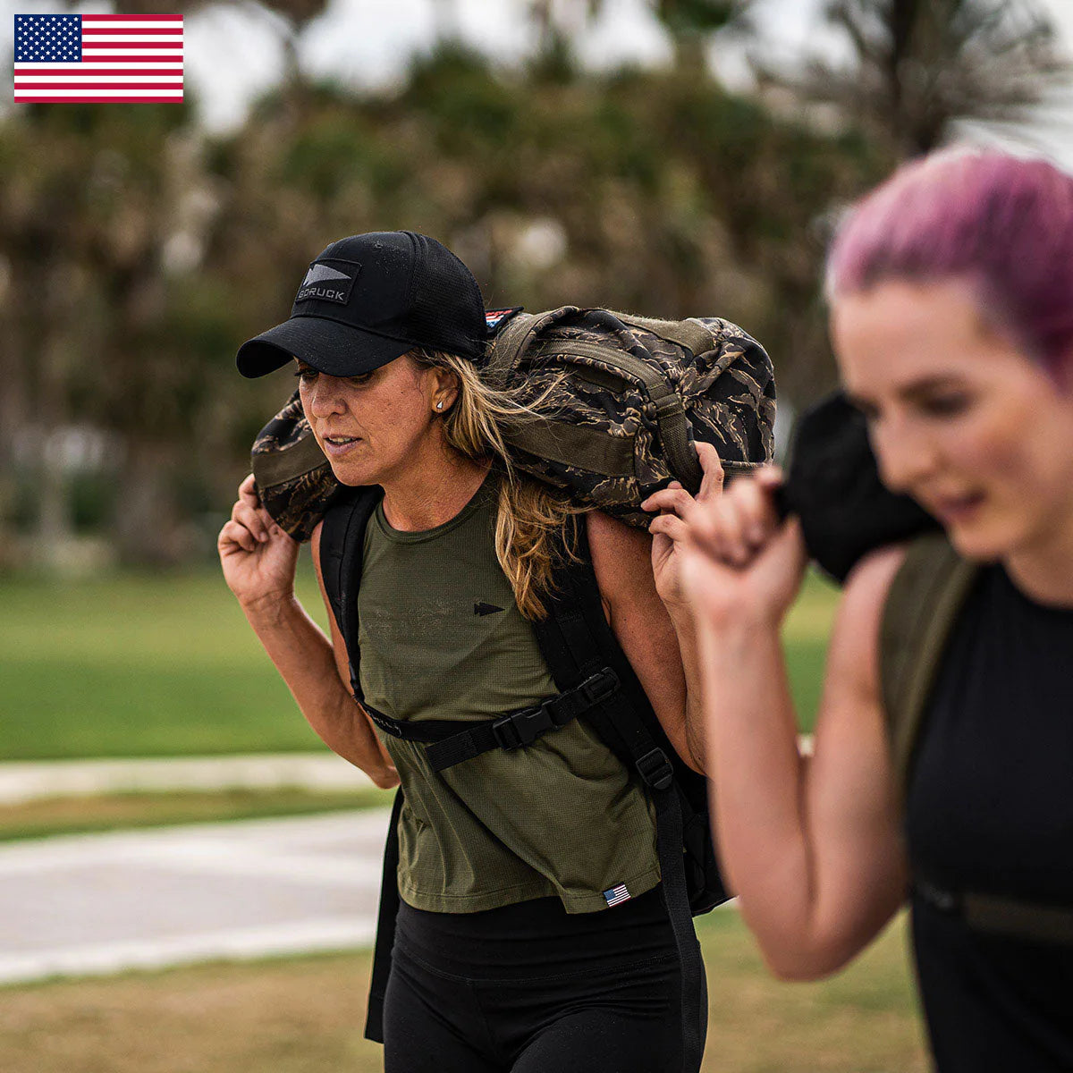 Two women outdoors carry heavy sandbags on their shoulders, both wearing the Women’s Performance Tank - ToughMesh, with an American flag in the corner.