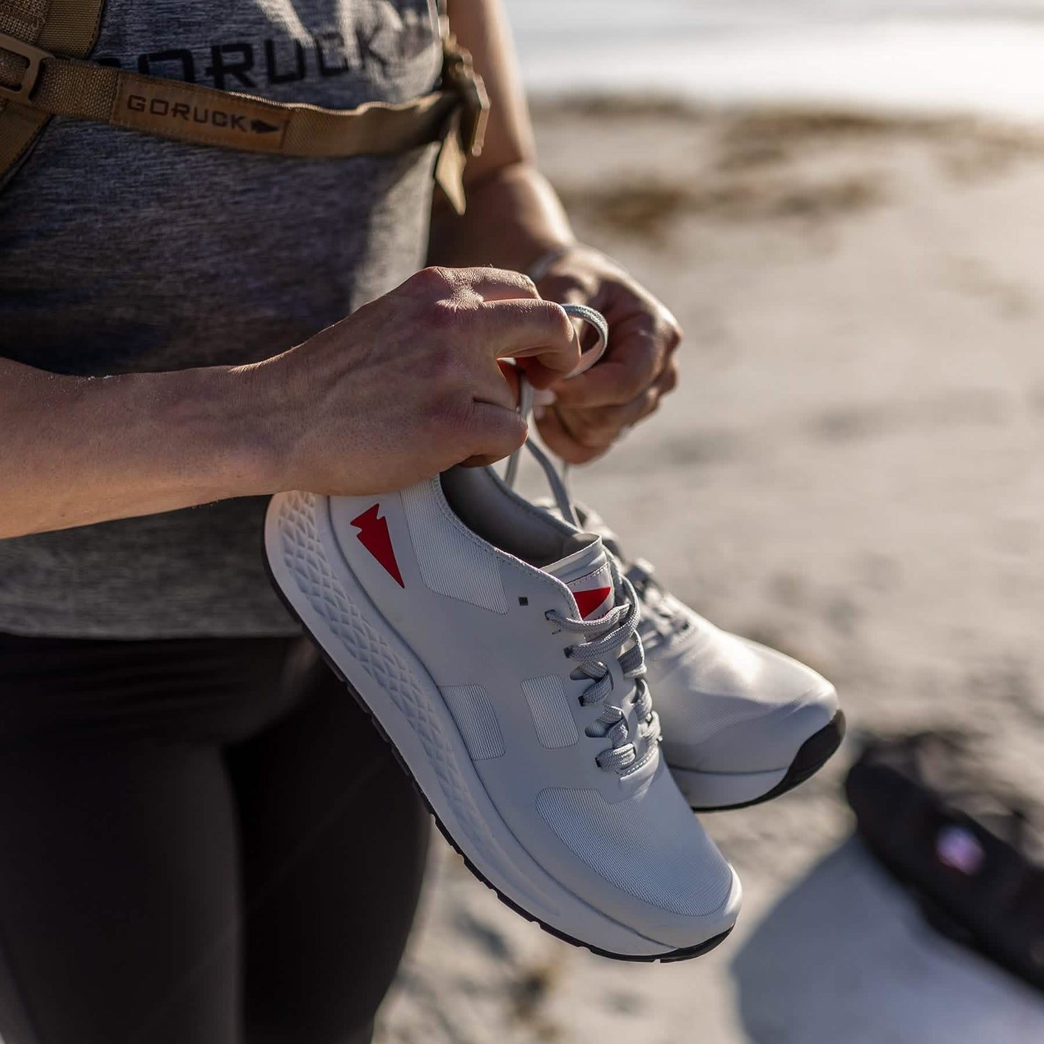 A person in a sleeveless top holds a pair of GORUCK Women's Rough Runner athletic shoes in light grey with red accents. The backdrop, featuring a sandy and sunlit area, suggests an outdoor setting perfect for high mileage running.