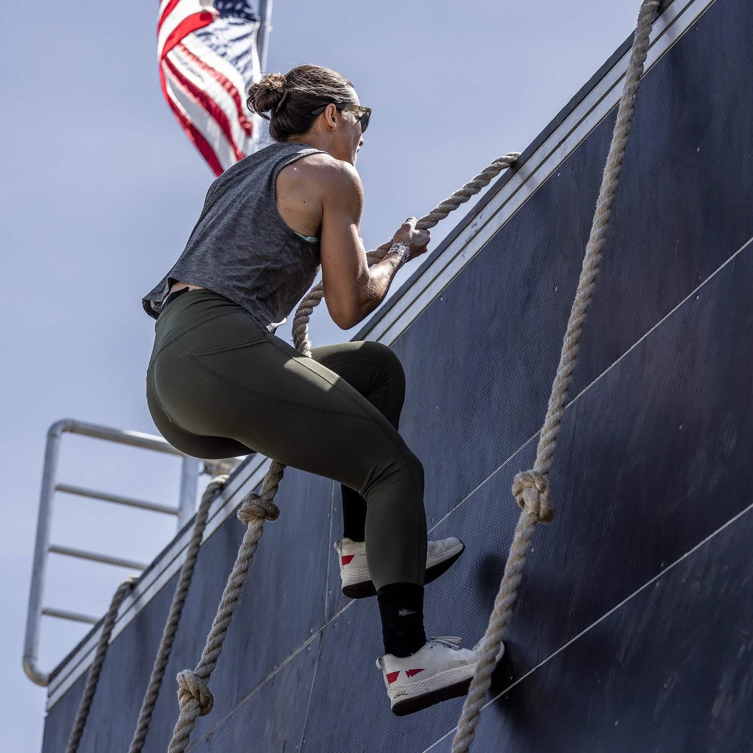 A woman dressed in athletic gear takes on a rope climb against a towering wall, with the American flag in the backdrop. She's sporting the GORUCK Women's Rough Runner in Light Grey and Red, paired with green leggings and high mileage running shoes. The clear blue sky provides perfect conditions for this Rough Runner challenge.