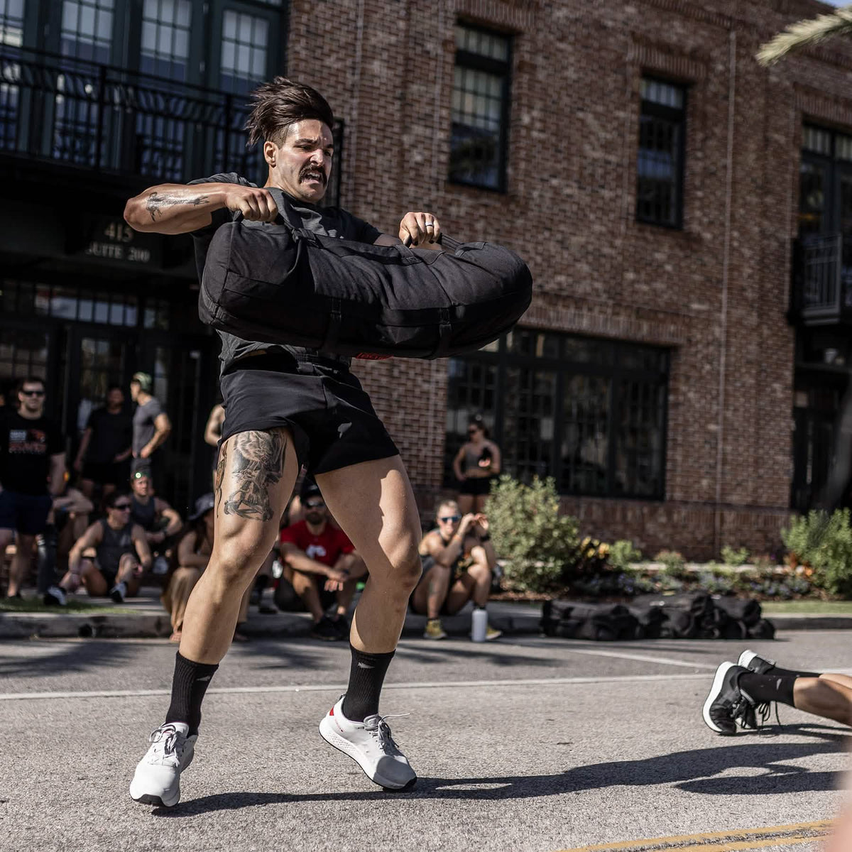 A man lifts a heavy sandbag outdoors at the Rough Runner event, showcasing his strength and endurance while wearing the Men's Rough Runner - Light Grey + Red shoes as spectators watch in the background.
