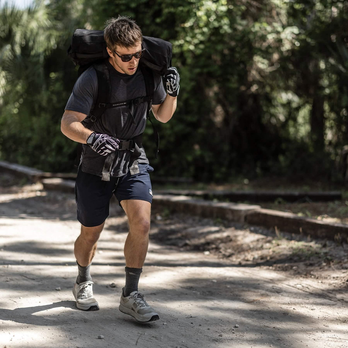 A man in sunglasses and gloves carries a large backpack along a sunny dirt path lined with trees, wearing Men's Rough Runner - Light Grey + Red shoes featuring a Gradient Density EVA midsole for added comfort.