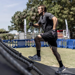 A man climbs an inclined rope outdoors in Men's Rough Runner - Light Grey + Red shoes, sporting athletic gear and gloves, with trees in the background.