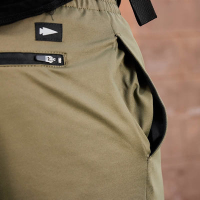 Close-up of GORUCK Men’s Performance Joggers made from high-quality performance fabric, showcasing an open pocket and a small black zipper pocket above it, adorned with a black tag displaying a white arrow symbol. The backdrop features a softly blurred wall in a warm brown hue.