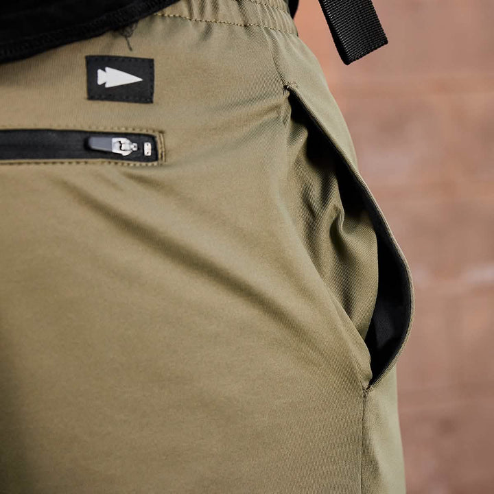 Close-up of GORUCK Men’s Performance Joggers made from high-quality performance fabric, showcasing an open pocket and a small black zipper pocket above it, adorned with a black tag displaying a white arrow symbol. The backdrop features a softly blurred wall in a warm brown hue.