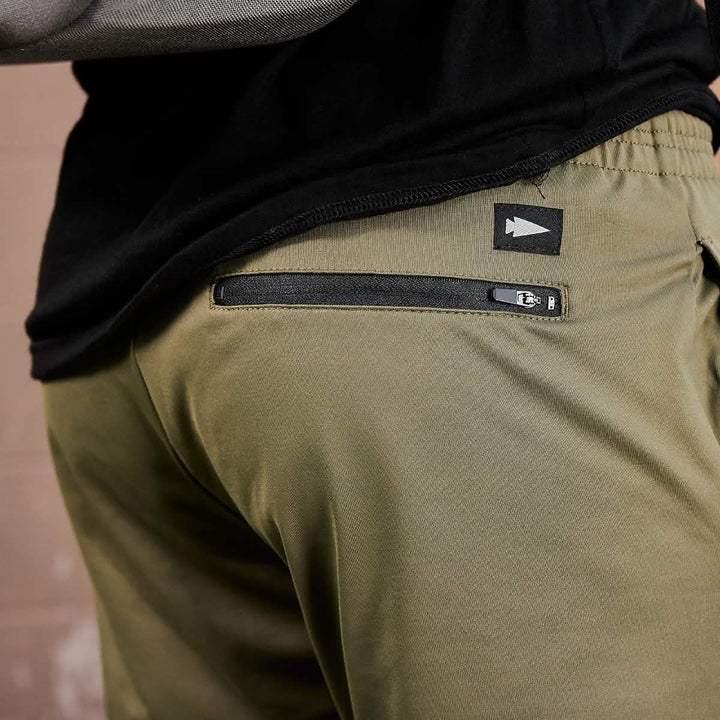 A close-up shows someone wearing GORUCK's Men’s Performance Joggers in olive green, showcasing a back zipper pocket. Made from moisture-wicking performance fabric, these joggers include a small black tag with a white arrow symbol, perfectly paired with a black shirt for a sleek appearance.
