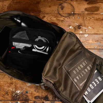 An open olive green Bullet Ruck - Classic - Reflective Spearhead by GORUCK rests on the wooden floor, displaying a stainless steel water bottle, a black mesh case with cables, a pen, and a partially visible book titled FREEDOM. This daypack is crafted with Cordura fabric and includes a mesh pocket on the flap.