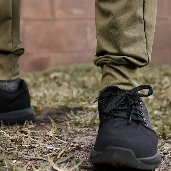 A close-up features a person sporting GORUCK's Men’s Performance Joggers in olive green, paired with dark sneakers, standing on grassy and slightly muddy terrain with a brick wall in the background.