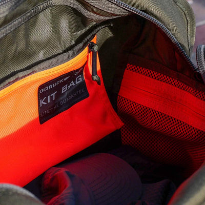 A close-up of an open green GORUCK Kit Bag (Includes Shoulder Strap), reminiscent of a Special Forces Flight Bag, reveals its orange interior lining and black tag labeled under the GORUCK brand with a Lifetime Guarantee. The interior features a mesh pocket and part of a cap, making it ideal for deployment readiness.