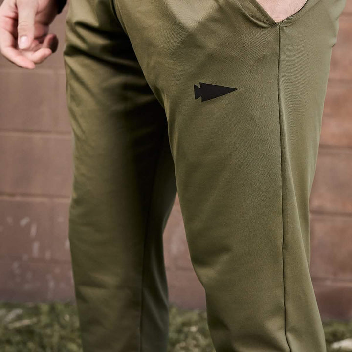 Close-up of a person wearing GORUCK Men’s Performance Joggers in olive green, showcasing a black arrowhead logo on the thigh. The moisture-wicking fabric provides comfort as the person's hand is slightly visible against a blurred outdoor backdrop with a brown brick wall and green grass.