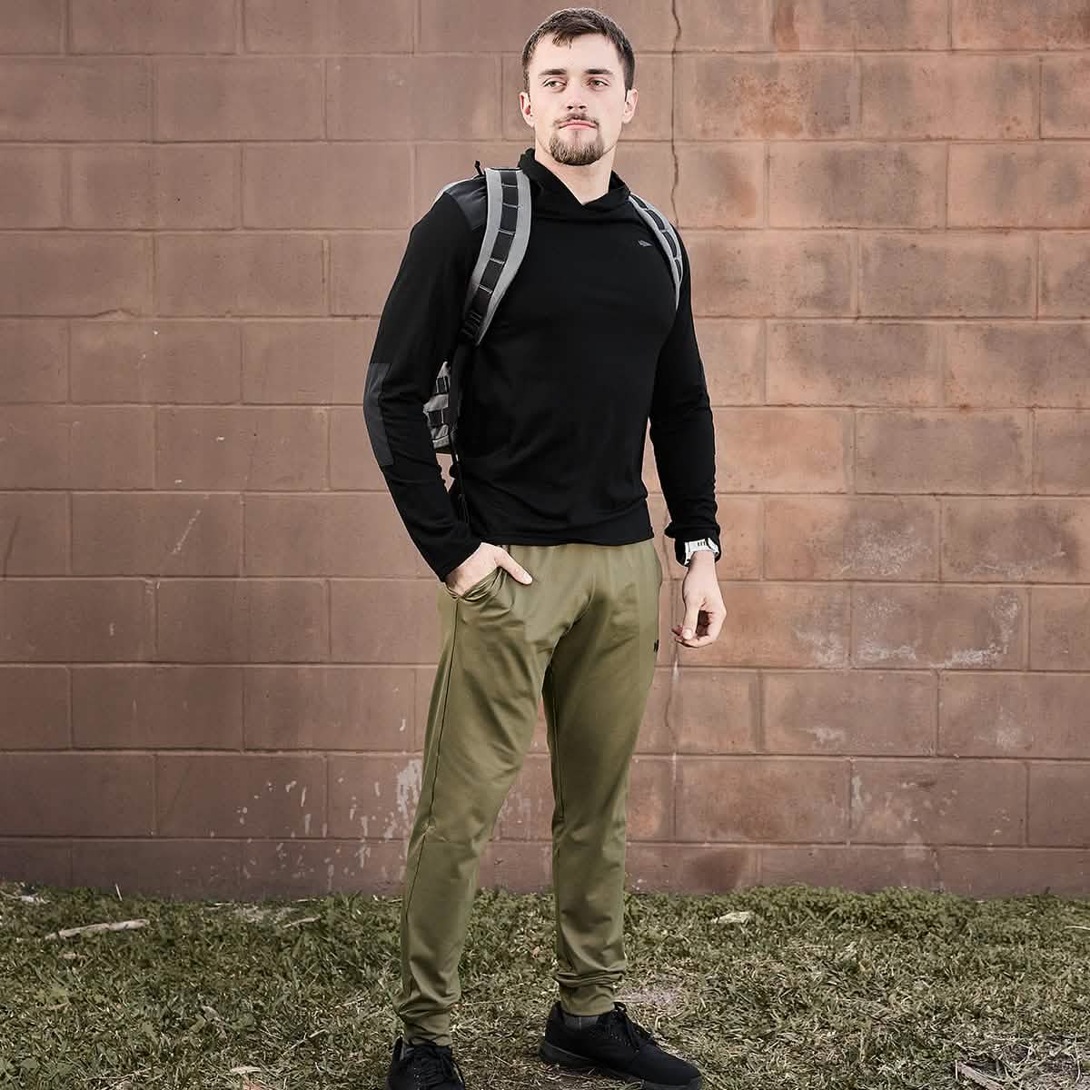 A person with short hair, dressed in a black hoodie and GORUCK's Men’s Performance Joggers in olive green, stands outdoors against a brick wall. They carry a gray backpack and have one hand in their pocket, standing on grass. The joggers' moisture-wicking fabric from GORUCK ensures comfort during any adventure.