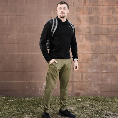 A person with short hair, dressed in a black hoodie and GORUCK's Men’s Performance Joggers in olive green, stands outdoors against a brick wall. They carry a gray backpack and have one hand in their pocket, standing on grass. The joggers' moisture-wicking fabric from GORUCK ensures comfort during any adventure.