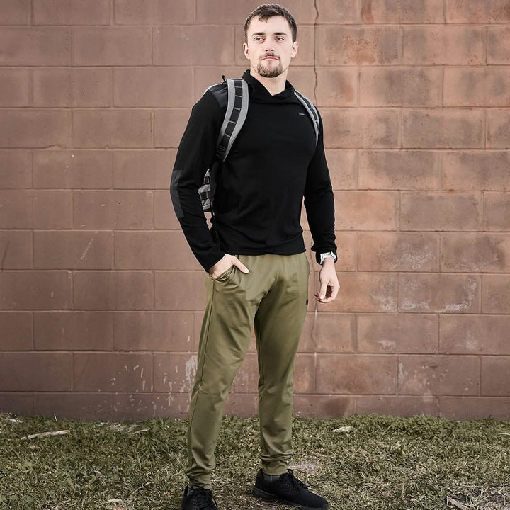 A person with short hair, dressed in a black hoodie and GORUCK's Men’s Performance Joggers in olive green, stands outdoors against a brick wall. They carry a gray backpack and have one hand in their pocket, standing on grass. The joggers' moisture-wicking fabric from GORUCK ensures comfort during any adventure.