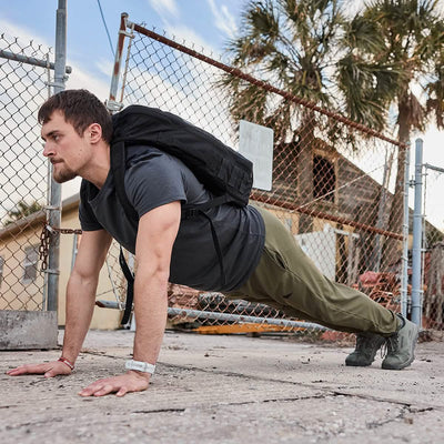 A person does a push-up on a concrete surface outdoors, wearing olive green GORUCK Men’s Performance Joggers, along with a black backpack and gray shoes. The moisture-wicking fabric of the joggers keeps them cool as they exercise near the chain-link fence, with palm trees and a brick building in the background.