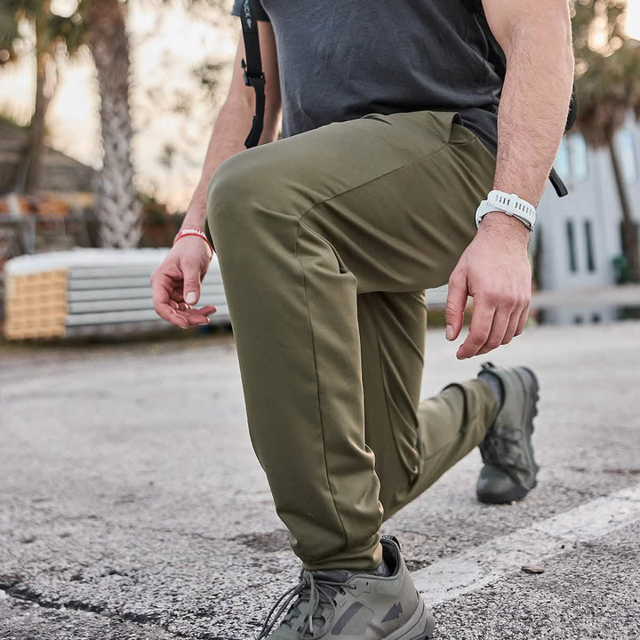A person wearing olive green GORUCK Men’s Performance Joggers, a dark gray shirt crafted from moisture-wicking performance fabric, and gray sneakers performs a lunge on an outdoor surface. They sport a white wristwatch and a red bracelet. Trees and a building are visible in the background.