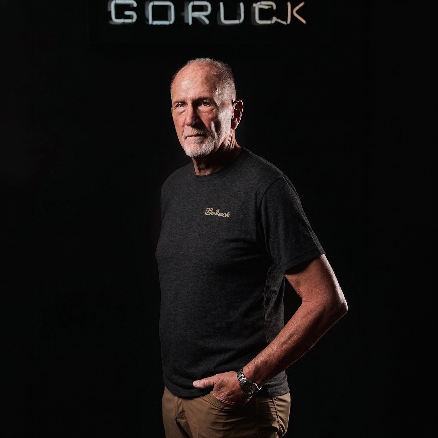An older man wears the GORUCK Bud Tee - Tri-BlendX, standing against a black background with a 
