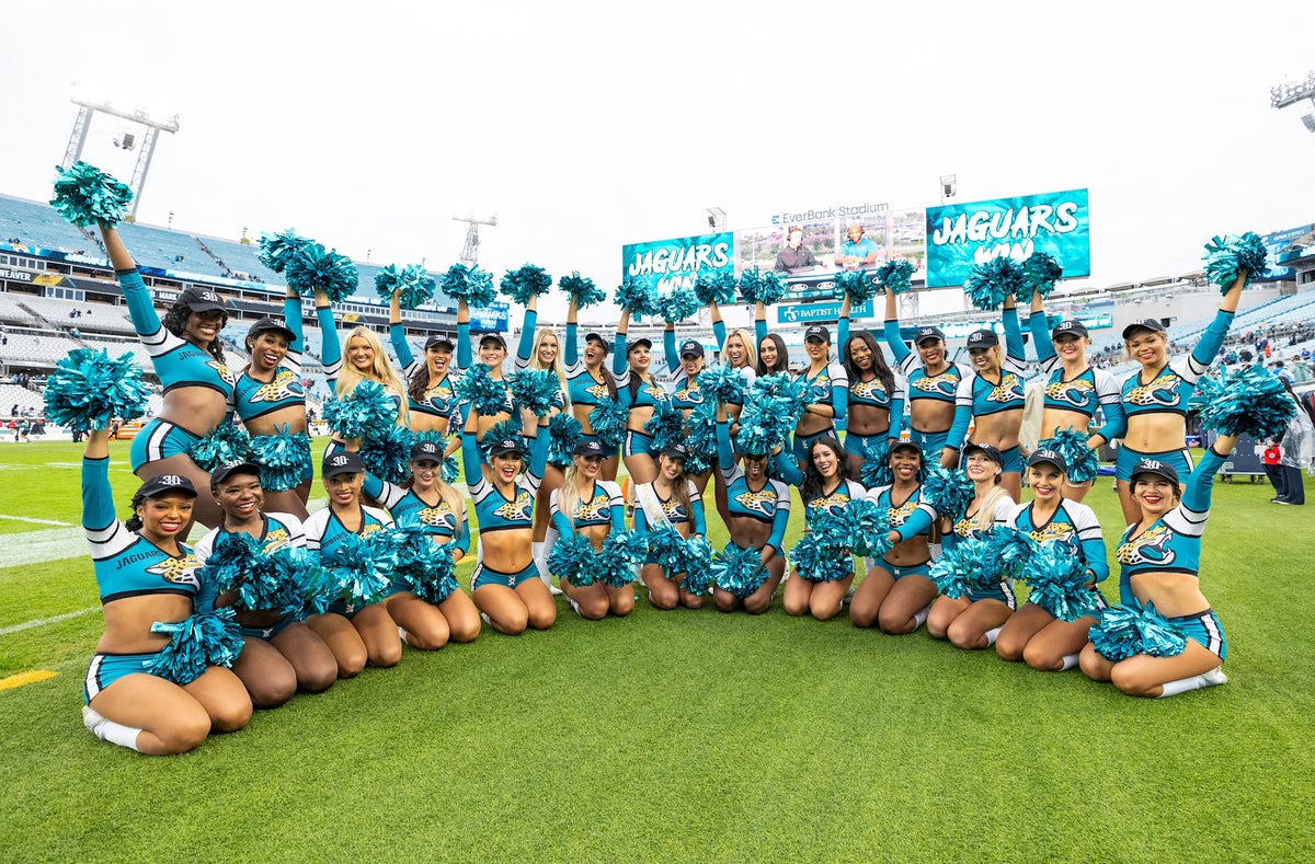 The Jacksonville Jaguars cheerleading squad in teal and black uniforms pose with teal pom-poms on the field at a Jacksonville Jaguars Foundation GORUCK event.