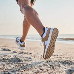 A low-angle shot captures a person running on a sandy beach, emphasizing their legs and the GORUCK Men's Rough Runner - Speed Tan sneakers designed for high mileage, with the ocean and horizon visible in the background under a clear sky.