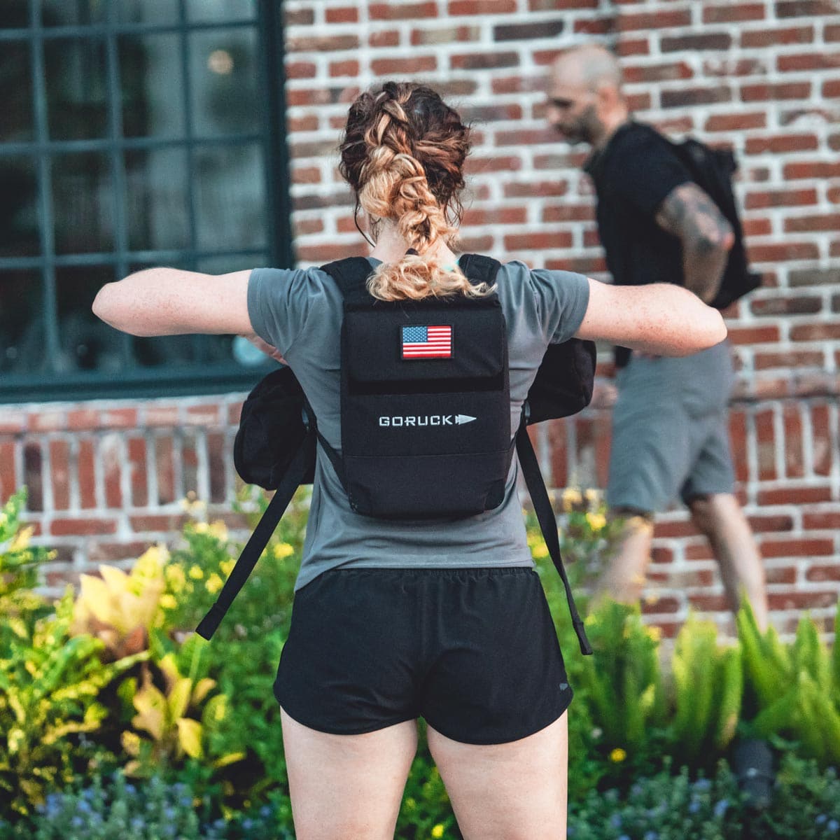 A woman with an American flag patch on her rucksack stands outside by a brick wall and greenery, ready for weighted training with her RPC 3.0 + 10lb Ruck Sandplate.