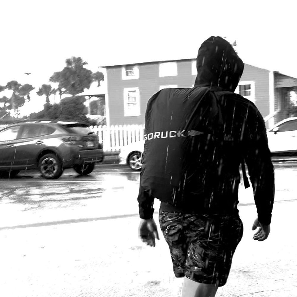 Person in a hooded jacket walking in the rain on a street, cars and houses in the background.