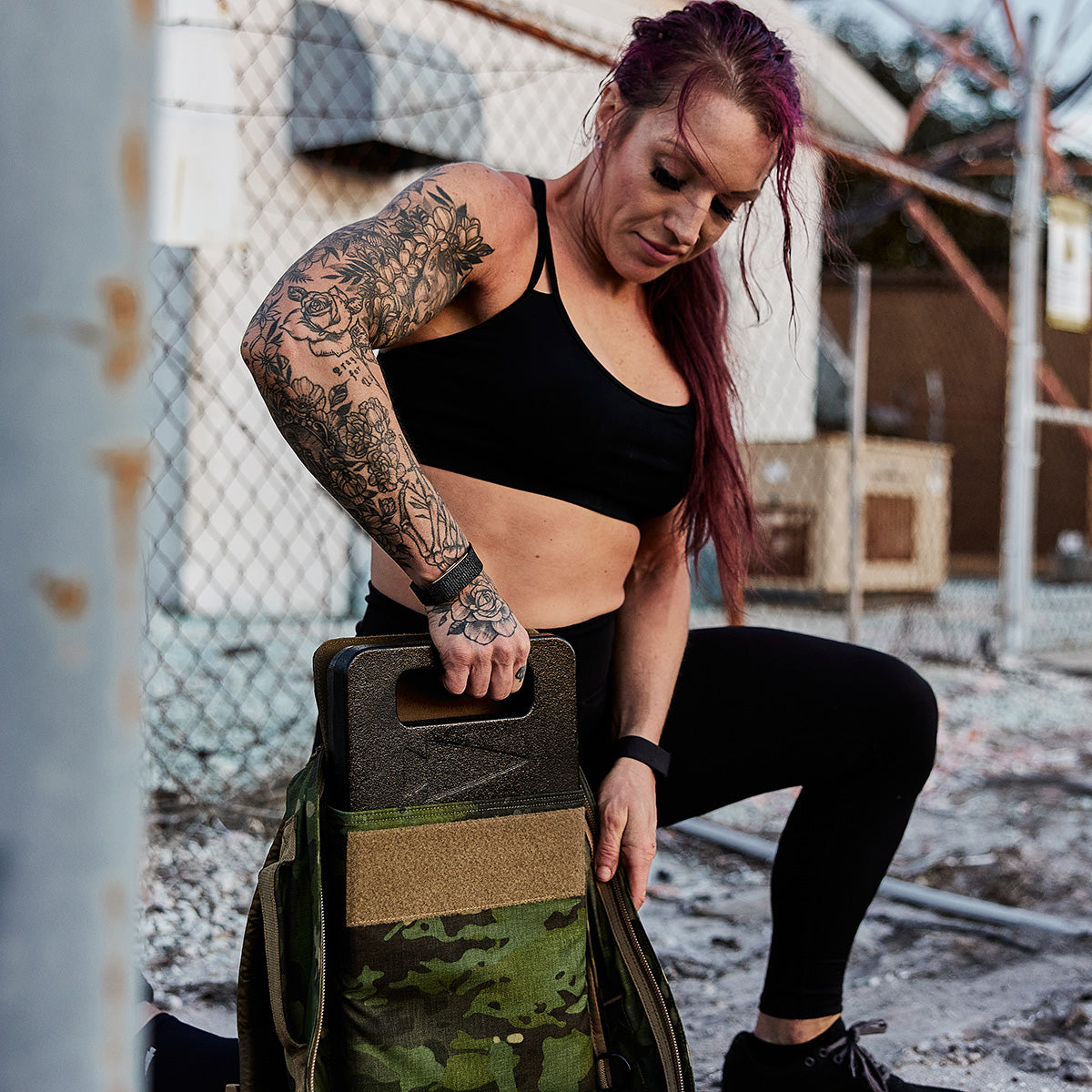 A fit, tattooed woman places a Ruck Plate into a camo backpack outdoors near a fence.