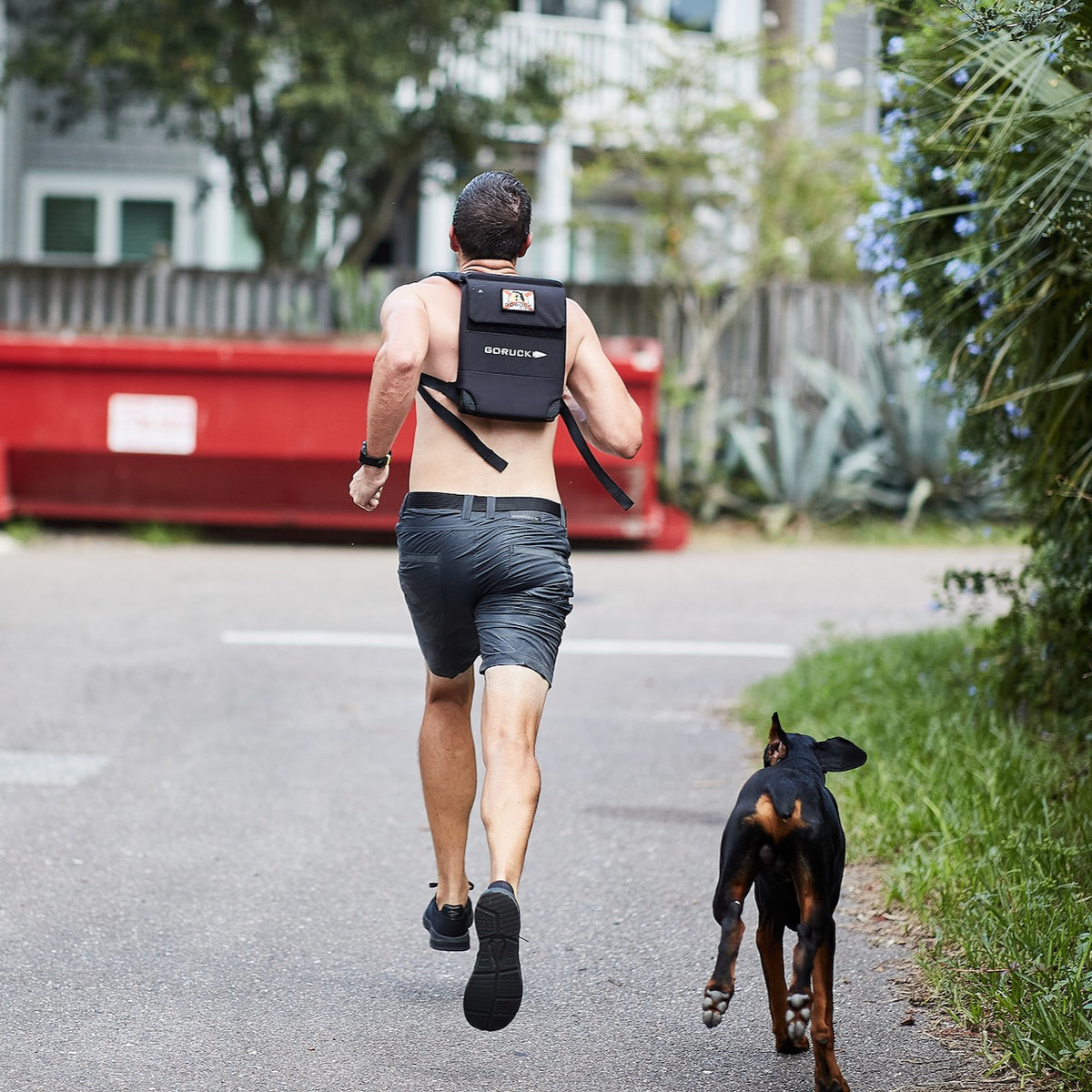 A shirtless man runs with a backpack down the street, followed by a black dog, showcasing the Spearhead Nylon Webbing Belt—an adjustable belt designed for both comfort and durability.