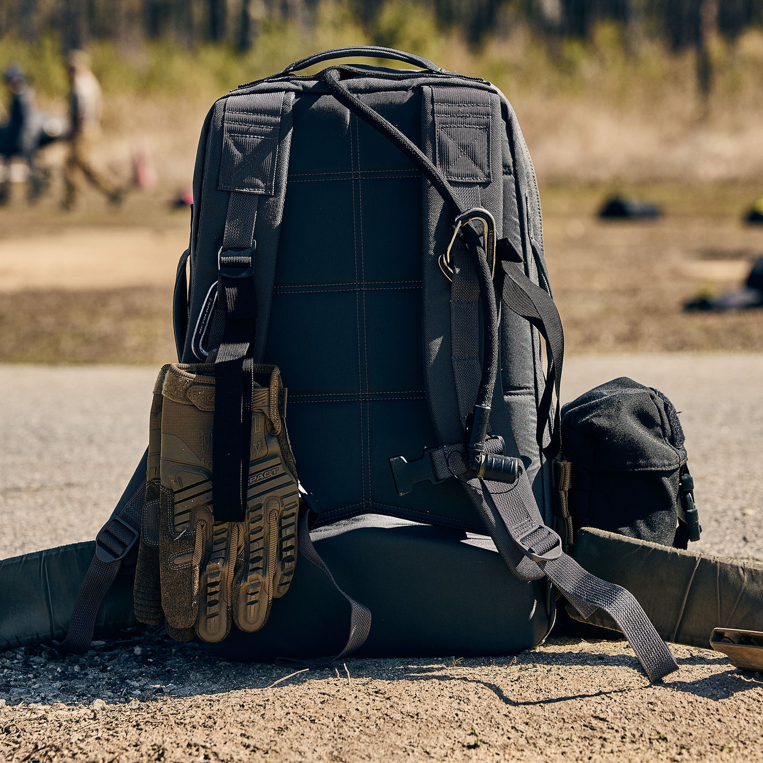 The Rucker 5.0, a black tactical rucksack with gloves and gear attached, sits outdoors on the ground, ready for any adventure and designed to provide reliable lumbar support.