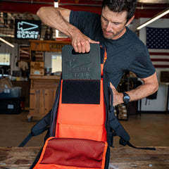 A man places a weighted plate into the Rucker 5.0, an orange and black rucksack designed for rucking, in a workshop with an American flag in the background.