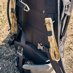 Close-up of the Rucker 5.0, a black rucksack with camouflage straps designed for rucking, resting on dry grass outdoors.