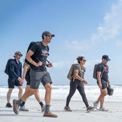 Five people with rucksacks walk together on a sandy beach under a blue sky with scattered clouds, enjoying a day of rucking and the comfort of the Rucker 5.0’s built-in lumbar support.