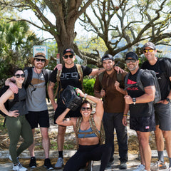 Seven people in athletic gear smile outdoors with Rucker 5.0 rucksacks, featuring lumbar support, and trees in the background.