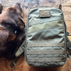 A brown dog lies next to an olive green Rucker 5.0 rucksack featuring built-in lumbar support on a wooden surface.