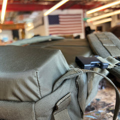 Close-up of the olive green Rucker 5.0 rucksack with lumbar support, with an American flag hanging in the blurred background.