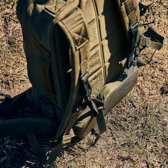 A close-up of the green Rucker 5.0 with straps and lumbar support, lying on dry grass outdoors—perfect for comfortable rucking adventures.