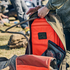 Outdoors with gear in the background, a person opens the Rucker 5.0, revealing its bright orange interior and DFQ patch—perfect for rucking adventures that require comfort and dependable lumbar support.