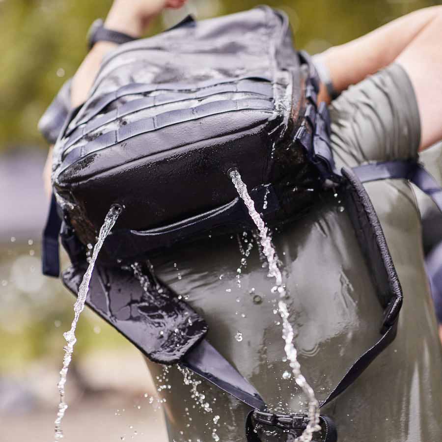 A wet backpack with water pouring out, being lifted by a person wearing a green shirt.