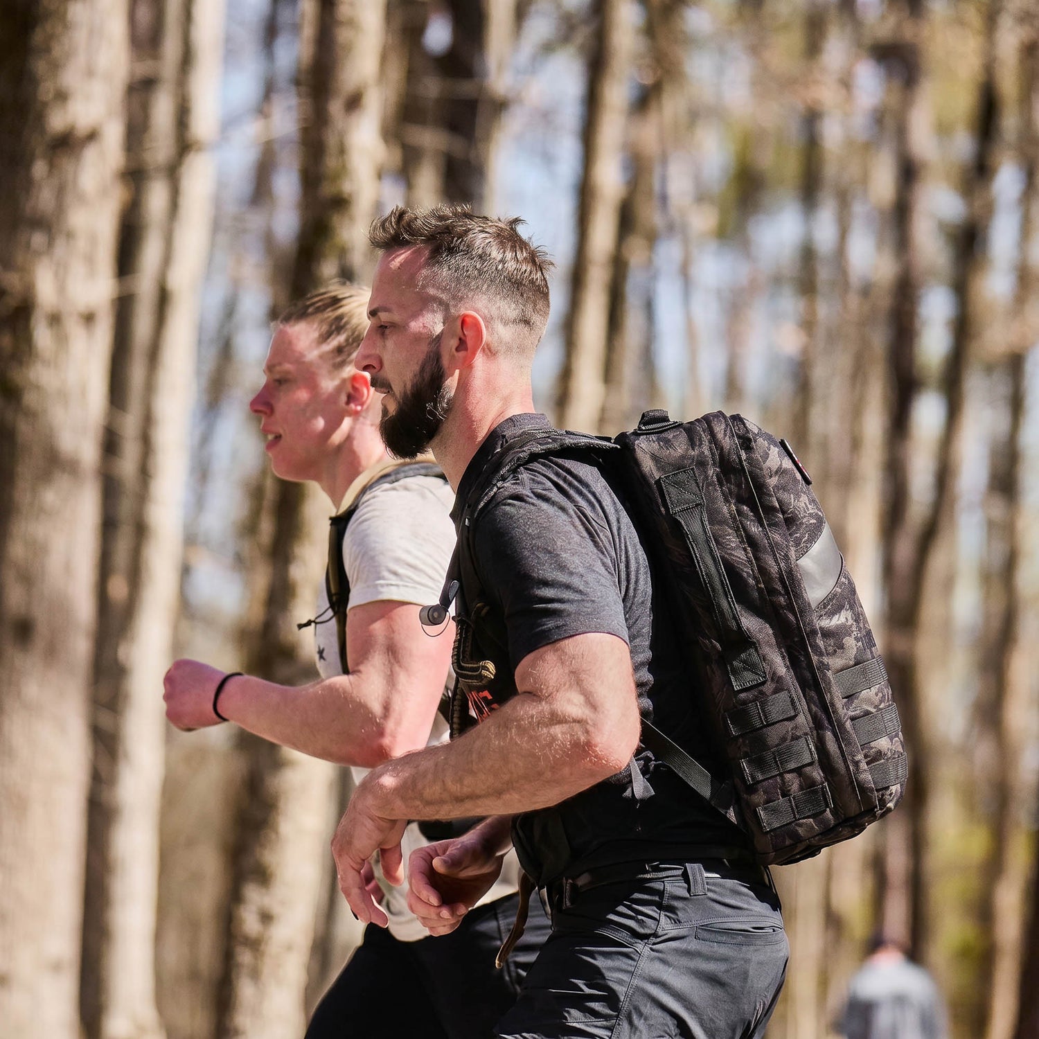 Two men with Rucker 5.0 packs run through a sunny, forested area, enjoying rucking and the extra lumbar support their gear provides.