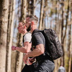 Two men with Rucker 5.0 packs run through a sunny, forested area, enjoying rucking and the extra lumbar support their gear provides.