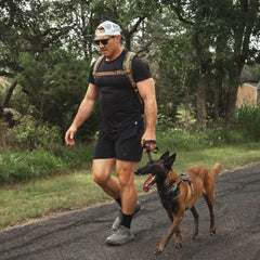 Man wearing a black outfit and backpack walking a brown dog on a leash outdoors on a paved path