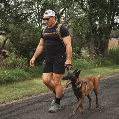 A man wearing a black t-shirt and shorts, paired with a white cap, confidently walks on the road with his brown dog on a leash. He sports sunglasses and carries a backpack while striding in his GORUCK Mackall - Earth + Forged Iron trail shoes, as trees and grass line the roadside behind him.