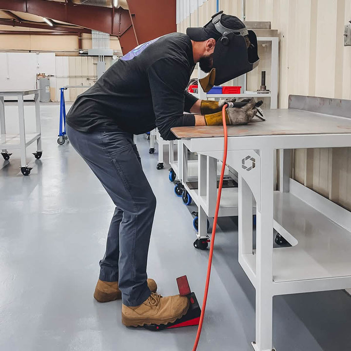 A person wearing a welding helmet, gloves, and the GORUCK MACV-2 Safety Boot - Mid Top is leaning over a metal table in an industrial workshop, crafting with precision. The space is equipped with mobile tables and various tools, meeting Special Forces standards for safety and efficiency.