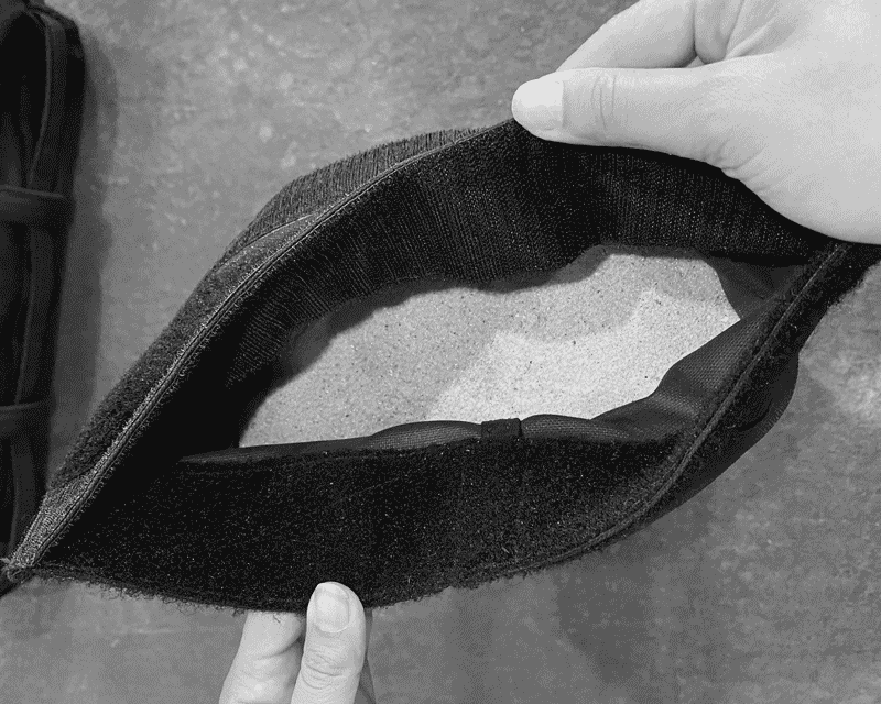 Hands holding open a black fabric pouch filled with sand, showing the contents inside.