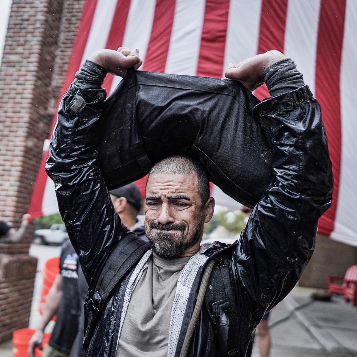 A man lifts a heavy sandbag overhead, with a large American flag hanging in the background.