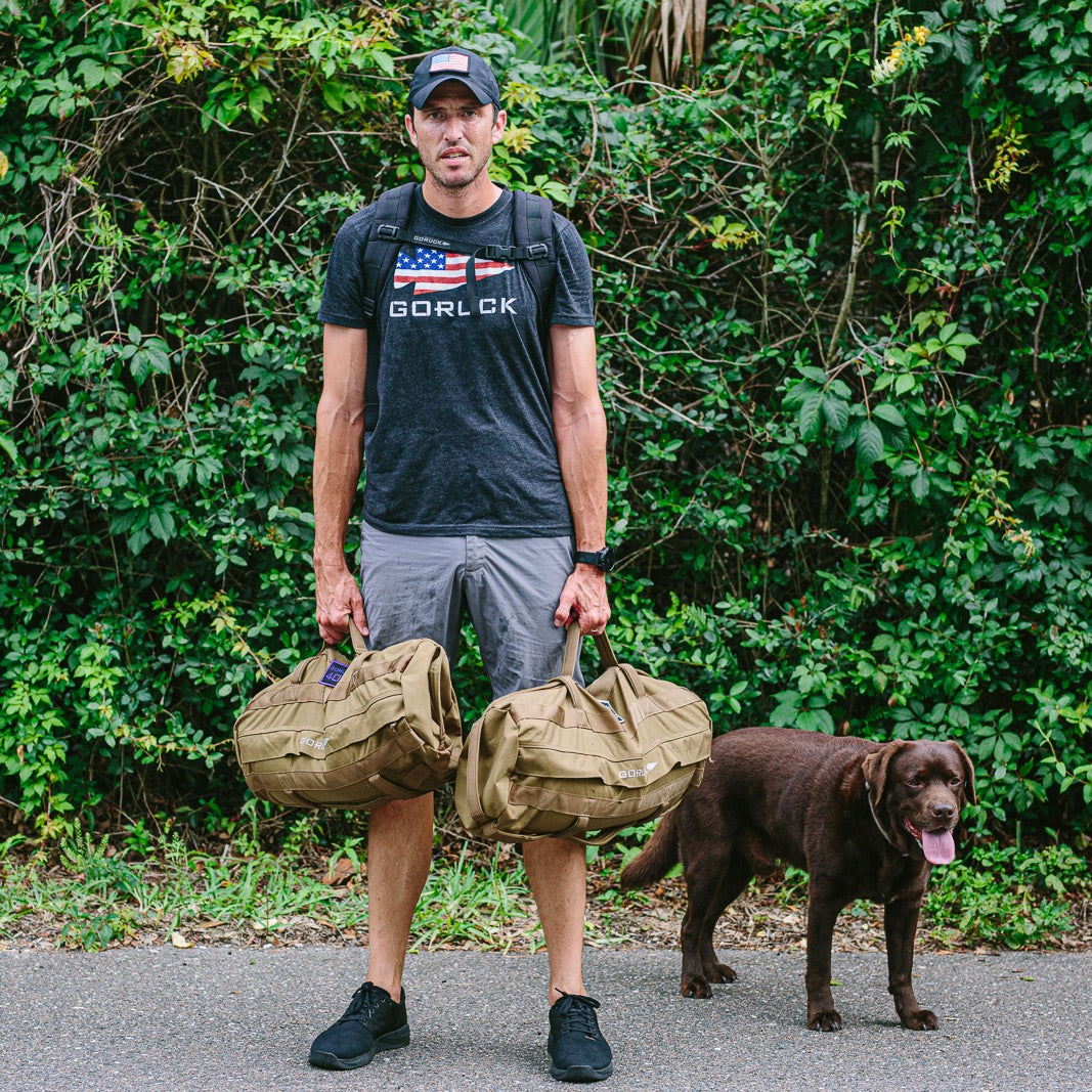 A man in a GORUCK t-shirt, hat, and Merino wool socks stands on a path holding two tan duffel bags, with a chocolate Labrador standing beside him.