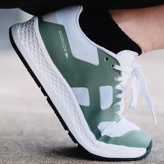 A close-up shot of a foot sporting the Women's Rough Runner - Seaspray by GORUCK, specifically designed for high mileage running. The shoe showcases a stylish green and white design with black laces and a textured white sole, set against concrete. Its Gradient Density EVA Midsole offers exceptional comfort and durability, making it perfect for conquering any Rough Runner challenge.