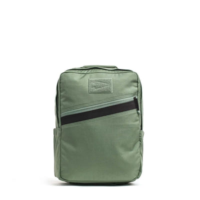 The KR1 by GORUCK is a green kids rucksack with a rectangular shape, featuring a black diagonal zipper pocket on the front. It includes a top handle, padded straps, and side pockets. Its simple design makes it suitable for both school supplies or professional use.