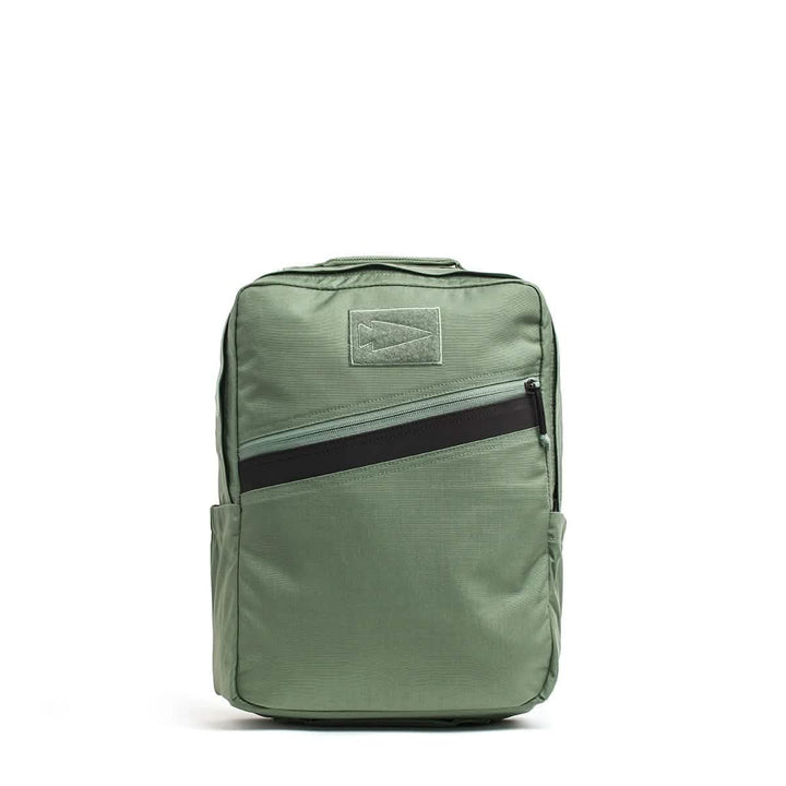The KR1 by GORUCK is a green kids rucksack with a rectangular shape, featuring a black diagonal zipper pocket on the front. It includes a top handle, padded straps, and side pockets. Its simple design makes it suitable for both school supplies or professional use.
